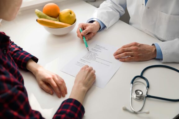 Doctor and patient discussing a treatment plan at a desk with a stethoscope and fruit bowl.