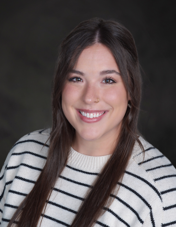 Smiling woman with long dark hair wearing a white knit sweater with black stripes, studio portrait against a dark background.